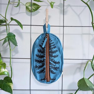 Pu&ograve; includere: Una pochette in tessuto blu di forma ovale con un disegno di un albero sempreverde stampato in nero. La pochette presenta una chiusura centrale verticale con cerniera marrone, dotata di una linguetta in tessuto e di un occhiello per appenderla.