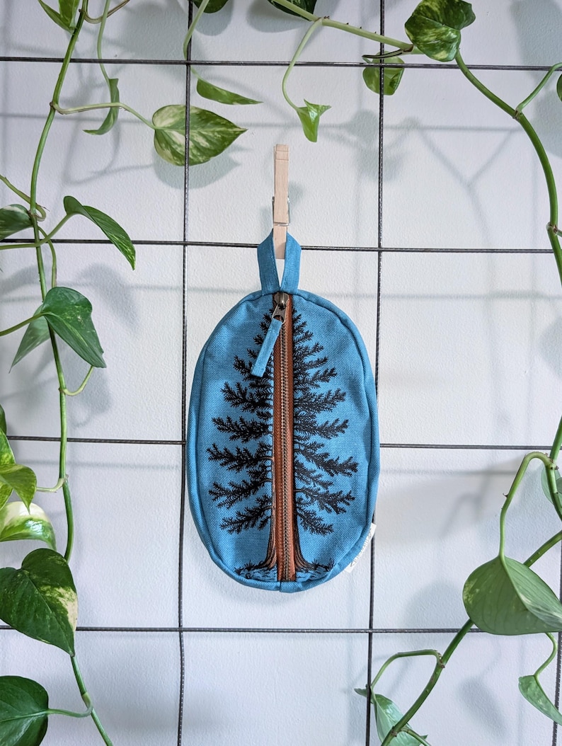 Pu&ograve; includere: Una pochette in tessuto blu di forma ovale con un disegno di un albero sempreverde stampato in nero. La pochette presenta una chiusura centrale verticale con cerniera marrone, dotata di una linguetta in tessuto e di un occhiello per appenderla.