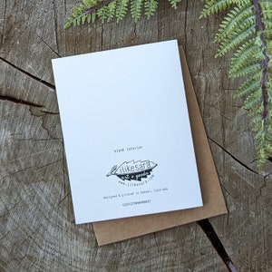 Op de afbeelding: Een witte wenskaart met de tekst "blank interior" en een logo voor "ilikesara art + goods" ligt op een bruine envelop. De kaart en envelop liggen op een rustiek houten oppervlak met groene varenaccenten.