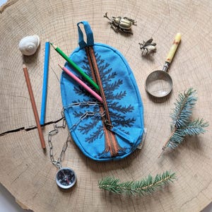 Pu&ograve; includere: Un astuccio ovale in tela blu brillante con stampa di pino nero e cerniera marrone. Contiene una matita verde e una rosa. Gli accessori includono una catenella argentata con bussola, una lente d'ingrandimento, una conchiglia e due insetti in metallo.