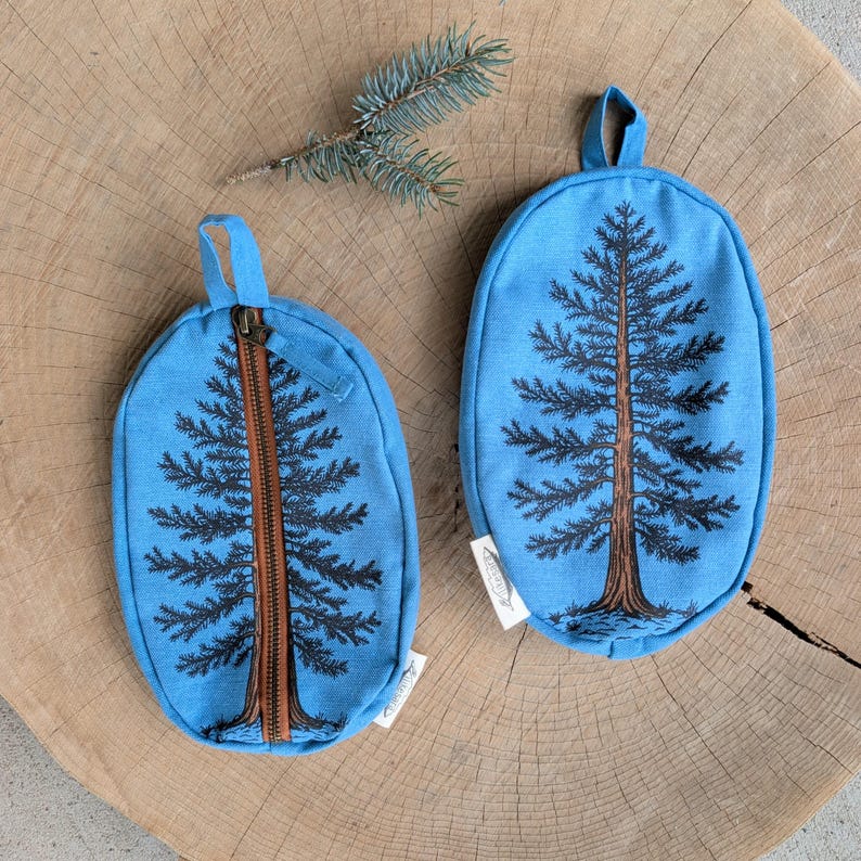 Pu&ograve; includere: Due pochette ovali in tessuto blu con un disegno di albero sempreverde stampato in nero e marrone. Una pochette ha una chiusura a zip verticale centrale, mentre l'altra &egrave; un pezzo unico con un occhiello per appenderla. Un rametto di pino &egrave; sopra.