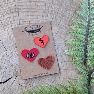 May include: A set of three iron-on patches featuring hearts with different designs. The hearts are red, orange, and black. The text on the card reads "iron-on patch set".