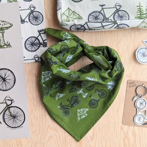 Pu&ograve; includere: Bandana verde con un motivo di bicicletta bianca. La bandana &egrave; piegata a forma di triangolo e ha un bordo bianco.