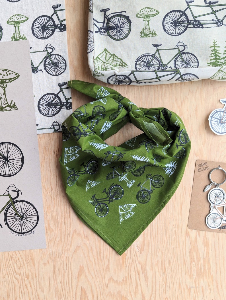 Pu&ograve; includere: Bandana verde con un motivo di bicicletta bianca. La bandana &egrave; piegata a forma di triangolo e ha un bordo bianco.