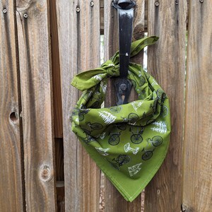 Pu&ograve; includere: Un bandana verde con un motivo di bicicletta e albero bianco. Il bandana &egrave; legato attorno a un chiavistello di cancello in metallo nero.