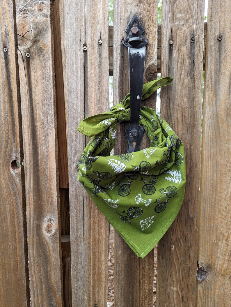 Pu&ograve; includere: Un bandana verde con un motivo di bicicletta e albero bianco. Il bandana &egrave; legato attorno a un chiavistello di cancello in metallo nero.