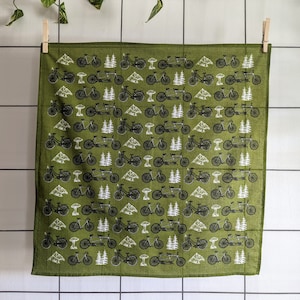 Pu&ograve; includere: Un bandana di stoffa verde con un motivo ripetitivo di biciclette nere e alberi. Il bandana &egrave; piegato e appeso a uno stendino.