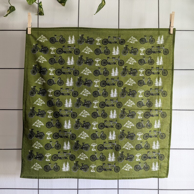 Pu&ograve; includere: Un bandana di stoffa verde con un motivo ripetitivo di biciclette nere e alberi. Il bandana &egrave; piegato e appeso a uno stendino.