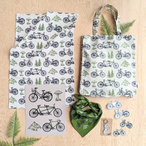 Pu&ograve; includere: Un set di quattro articoli con un design a tema bicicletta. Il set include una borsa di tela verde e bianca, un asciugamano da cucina verde e bianco, una bandana verde e un set di quattro adesivi per bicicletta verde e bianco. La borsa di tela ha uno sfondo bianco con biciclette verdi e foglie verdi. L'asciugamano da cucina ha uno sfondo bianco con biciclette verdi e foglie verdi. La bandana ha uno sfondo verde con biciclette bianche e foglie bianche. Gli adesivi hanno uno sfondo bianco con biciclette verdi e foglie verdi.