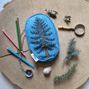 Pu&ograve; includere: Una pochette ovale in tessuto azzurro con un'illustrazione di un pino in nero e marrone. La pochette presenta una piccola etichetta bianca sul lato. &Egrave; circondata da matite colorate, una bussola argentata, una lente d'ingrandimento, una chiocciola e aghi di pino.