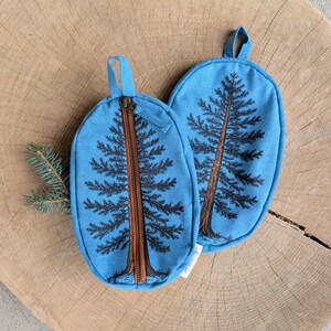 Pu&ograve; includere: Due pochette ovali in tessuto blu con un disegno di pino stampato in marrone scuro. Ogni pochette presenta un passante in tessuto sulla parte superiore e una cerniera verticale centrale con bordo marrone su una superficie in legno chiaro.
