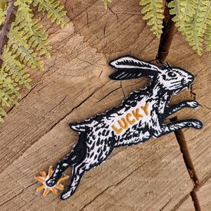 May include: Black and white embroidered patch of a running rabbit with the word "LUCKY" written across its body in yellow thread. The rabbit has a four-pointed star at the end of its paw.