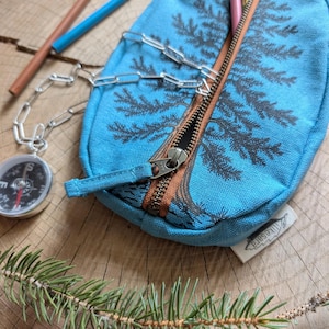 Pu&ograve; includere: Un astuccio in tela blu brillante con un disegno di pino serigrafato in grigio scuro. L'astuccio presenta una chiusura a zip marrone, una catena in metallo color argento e una piccola bussola attaccata. &Egrave; visibile un'etichetta in tessuto bianco con il testo likesara.