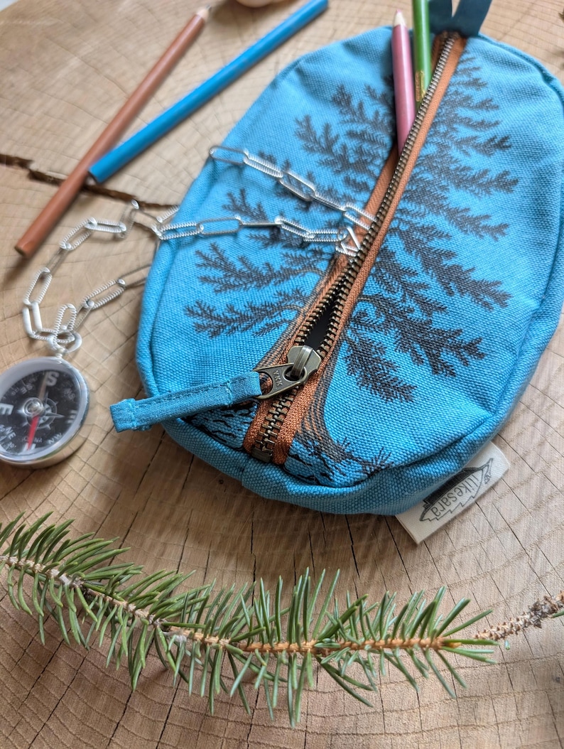 Pu&ograve; includere: Un astuccio in tela blu brillante con un disegno di pino serigrafato in grigio scuro. L'astuccio presenta una chiusura a zip marrone, una catena in metallo color argento e una piccola bussola attaccata. &Egrave; visibile un'etichetta in tessuto bianco con il testo likesara.