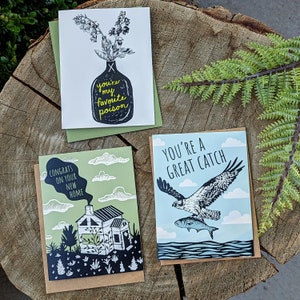 Op de afbeelding: Drie wenskaarten met illustraties en tekst. E&eacute;n kaart heeft een zwarte vaas met bloemen en de tekst "you're my favorite poison". Een andere kaart zegt "Congrats on your new home" met een huisafbeelding. De derde kaart zegt "You're a great catch" met een adelaar.
