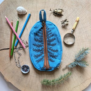 Pu&ograve; includere: Un astuccio ovale in tela blu brillante con una grafica di pino nero centrata su una cerniera marrone. L'astuccio &egrave; circondato da una conchiglia bianca, tre matite colorate, una bussola argentata, uno scarabeo metallico, una lente d'ingrandimento e aghi di pino.