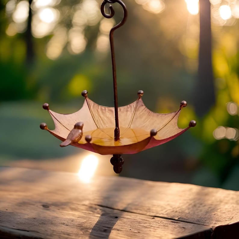 May include: A metal bird feeder shaped like an upside down umbrella with a small bird perched on the edge. The feeder is painted in shades of brown and yellow with a clear water basin. The feeder is hanging from a metal hook against a blurred background of green foliage and a bright orange sunset.
