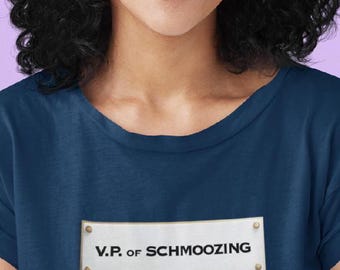 Puede incluir: Una camiseta azul marino con una etiqueta rectangular dorada que dice "V.P. of SCHMOOZING".
