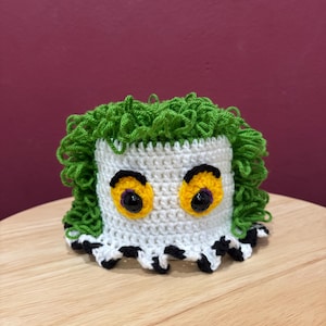 Könnte beinhalten: Gehäkelte Figur in Weiß und Grün mit großen gelben Augen und grünen Haaren. Die Figur hat einen schwarz-weißen Zierrand an der Basis. Das handgefertigte Objekt ist wahrscheinlich ein Dekorationsobjekt oder ein Spielzeug.