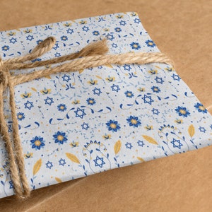 K&ouml;nnte beinhalten: Ein Geschenk, das in wei&szlig;es Papier mit einem blauen und gelben Blumenmuster mit Davidsternen eingewickelt ist. Das Geschenk ist mit braunem Garn gebunden.