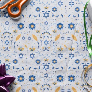K&ouml;nnte beinhalten: Wei&szlig;es Geschenkpapier mit einem blauen und gelben Blumenmuster und einem sich wiederholenden Davidstern-Design. Das Papier ist mit einem gr&uuml;nen Band, einem goldenen Band und einer lila Schleife verziert.