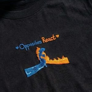 以下が含まれることがあります： 黒いTシャツ。刺繍されたテキストとグラフィックが特徴です。「Opposites React」の文字は青とオレンジで、ハートマークが付いています。グラフィックは青とオレンジの2つの図形です。カジュアルな普段着です。
