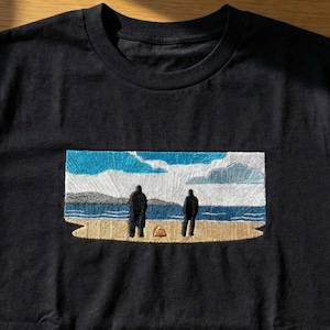 May include: Black crew neck t-shirt featuring an embroidered scene of two figures on a beach, with a blue sky and ocean backdrop. The design is detailed with various colors, creating a scenic view. The shirt is made of a soft material.