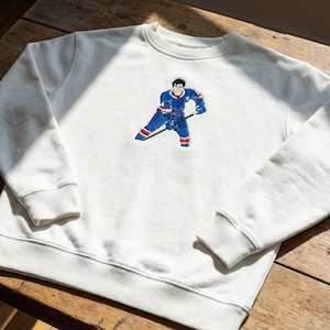 May include: Off-white sweatshirt with a hockey player graphic. The embroidered design shows a player in blue and red uniform, holding a hockey stick. The sweatshirt has a crew neck and ribbed cuffs.