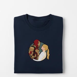 May include: Navy blue sweatshirt featuring an embroidered design of a superhero and a woman in a loving embrace. The superhero is in a red and gold suit. The woman has blonde hair and is wearing a white dress. The design is in a circular shape.