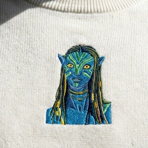 May include: A cream-colored crewneck sweatshirt featuring a detailed machine-embroidered design of a blue-skinned humanoid character with yellow eyes, pointed ears, and long dark hair with green highlights, centered on the chest area.