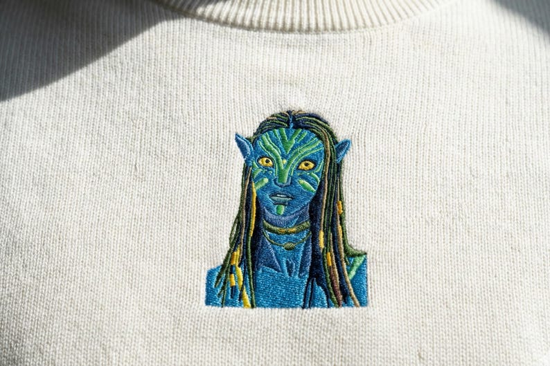 May include: A cream-colored crewneck sweatshirt featuring a detailed machine-embroidered design of a blue-skinned humanoid character with yellow eyes, pointed ears, and long dark hair with green highlights, centered on the chest area.
