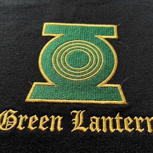 May include: A black fabric surface featuring an embroidered Green Lantern logo in green and gold thread. The circular emblem is centered above the text Green Lantern, which is stitched in a gold-colored, stylized serif font.