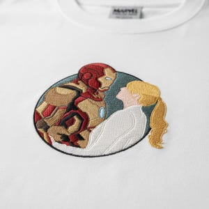 Peut inclure: Un t-shirt blanc avec un motif circulaire brodé représentant Iron Man en armure rouge et or face à une personne aux cheveux blonds en queue de cheval portant une chemise blanche. Le logo Marvel est imprimé sur le col intérieur du vêtement.