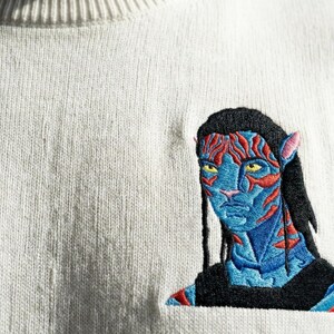 May include: A cream-colored knit sweater featuring a detailed embroidered patch of a blue-skinned humanoid character with yellow eyes, black hair, and red facial markings. The embroidery displays intricate stitching in shades of blue, red, and black.