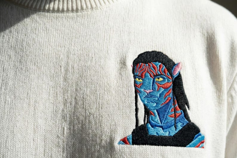 May include: A cream-colored knit sweater featuring a detailed embroidered patch of a blue-skinned humanoid character with yellow eyes, black hair, and red facial markings. The embroidery displays intricate stitching in shades of blue, red, and black.