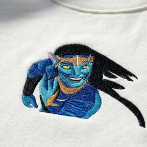 May include: Embroidered design of a blue Na'vi character from the movie Avatar on a white sweatshirt. The character has black hair, yellow eyes, and a gold headpiece. The design is detailed and colorful.
