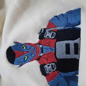 May include: An off-white fabric garment featuring a detailed embroidered patch of a blue-skinned character with yellow eyes, wearing a red and black suit with blue accents. The character has pointed ears and a black hairstyle, depicted from the chest up.