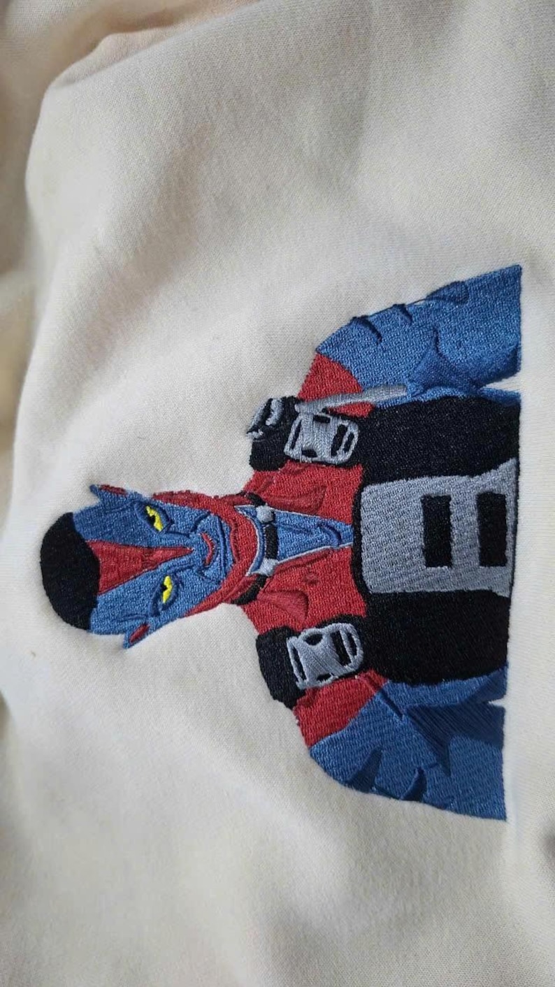 May include: An off-white fabric garment featuring a detailed embroidered patch of a blue-skinned character with yellow eyes, wearing a red and black suit with blue accents. The character has pointed ears and a black hairstyle, depicted from the chest up.