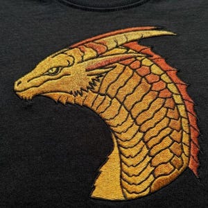 Può includere: Una t-shirt nera con una dettagliata testa di drago ricamata di profilo. Il drago è raffigurato in tonalità di giallo, oro e arancione, con texture a scaglie visibili e un corno prominente che si estende all'indietro dalla testa.
