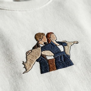 Puede incluir: Sudadera de color crema con un diseño bordado de dos figuras en un abrazo romántico, con los brazos extendidos. El bordado utiliza hilo marrón, beige y azul marino. La sudadera tiene cuello redondo acanalado.