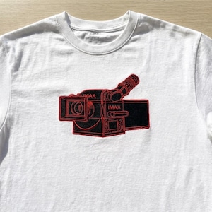 Könnte beinhalten: Ein weißes T-Shirt mit kurzen Ärmeln und einer schwarz-roten Stickerei einer professionellen Filmkamera. Das Kameramotiv zeigt den Schriftzug IMAX in roten Buchstaben an der Seite und oben sowie ein markantes Objektiv und einen Sucher.