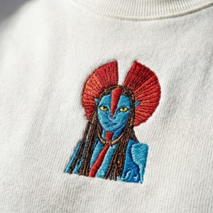 May include: An off-white sweatshirt featuring a detailed embroidered graphic of a blue-skinned humanoid character with yellow eyes, long dark hair, and a prominent red fan-shaped headpiece, centered on the chest area of the garment.