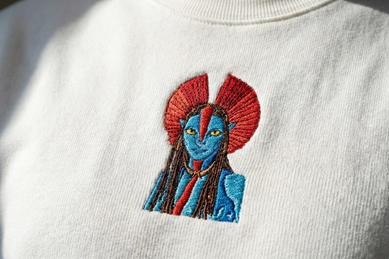 May include: An off-white sweatshirt featuring a detailed embroidered graphic of a blue-skinned humanoid character with yellow eyes, long dark hair, and a prominent red fan-shaped headpiece, centered on the chest area of the garment.