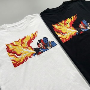 May include: Two t-shirts, one white and one black, each featuring a colorful embroidered design. The design depicts a fiery explosion with figures in the center. The figures are in shades of blue, red, and white.