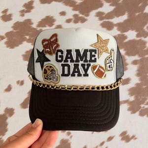 Op de afbeelding: Een wit met zwarte truckerpet met de tekst GAME DAY in zwarte letters. De pet is versierd met patches, waaronder een bruine strik, een gouden ster, een zwarte ster, een footballhelm met luipaardprint, een football en een schuimrubberen vinger. Een gouden ketting siert de zwarte klep.