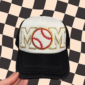 Puede incluir: Gorra de camionero blanca con una malla negra en la parte posterior. La parte delantera de la gorra presenta un MOM de purpurina dorada con una pelota de béisbol roja y blanca en el centro.