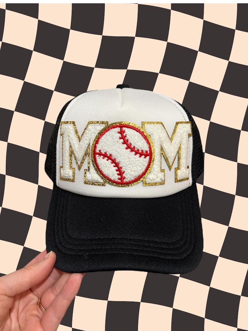 May include: White trucker hat with a black mesh back. The front of the hat features a gold glitter MOM with a red and white baseball in the center.
