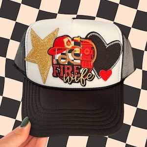May include: A black and white trucker hat with a white front panel featuring a gold star, a black heart, and a red heart. The hat has the words "FIRE WIFE" in gold script, and a firefighter's helmet graphic.