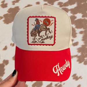 May include: A cream-colored baseball cap with a red brim. The front panel features a square embroidered patch depicting a cowboy riding a bucking horse with a red sun, and the word Cowboy. The red brim features the word Howdy in white script.