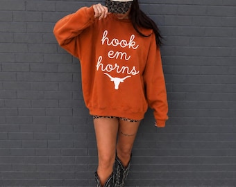 Pode incluir: Uma camisola oversize em laranja queimado com o texto branco hook em horns e um gráfico de boi longhorn. O conjunto inclui um boné de basebol creme com texto vermelho I LOVE YOU e aba com padrão leopardo, além de botas de cowboy pretas.